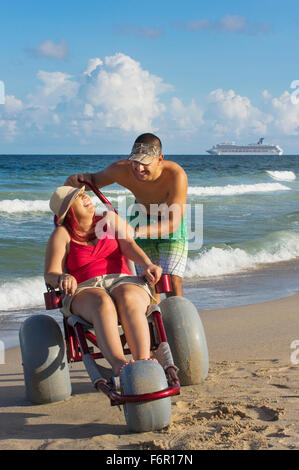 Menschen drängen Freundin querschnittsgelähmt im Rollstuhl am Strand Stockfoto