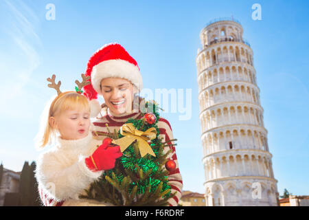 Glückliche Mutter in Weihnachtsmütze und Tochter tragen lustige Rentier-Geweih halten Weihnachtsbaum vor schiefen Tour von Pis Stockfoto
