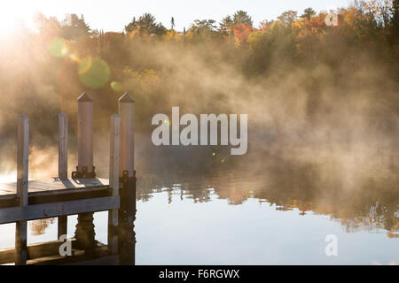 Morgennebel schwebt über einer ruhigen See mit Sonne und Lens Flare und ein Pier.  Viele der Stimmung im Bild mit führenden Linien zu Nebel. Stockfoto