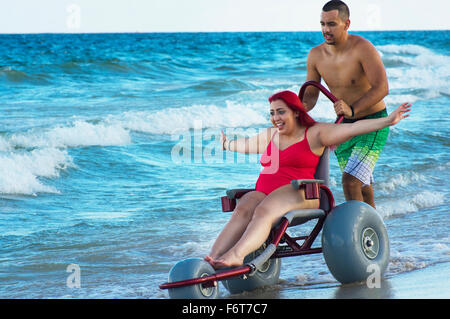 Menschen drängen Freundin querschnittsgelähmt im Rollstuhl am Strand Stockfoto