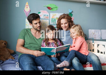 Paar und kleinen Töchter sitzen zusammen auf dem Bett im Kinderzimmer ein Buch zu lesen Stockfoto