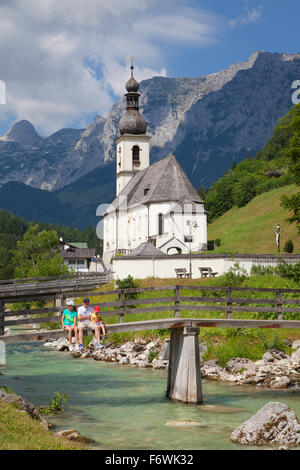 Vater und Kinder sitzen auf einem Fußgänger Brücke über die Ramsauer Ache vor Ramsau Kirche, Blick zur Reiteralpe, Berchtesgad Stockfoto