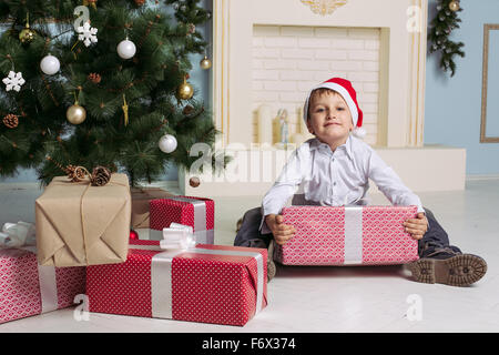 Kind in Nikolausmütze sitzt in der Nähe von Weihnachtsbaum mit Geschenk in Händen Stockfoto