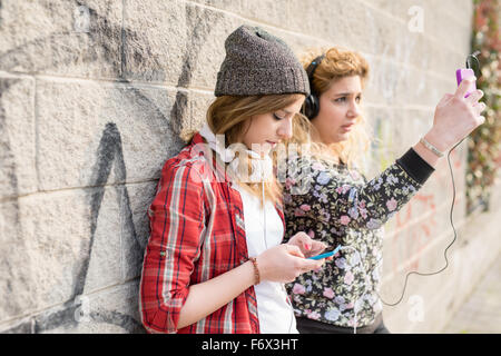 Halbe Länge von zwei jungen lockiges und glattes blondes Haar kaukasischen Frau eine Wand gelehnt, Musik hören und per Smartphone handheld, konzentrieren sich einerseits im Vordergrund - Technologie, Musik, Kommunikations-Konzept Stockfoto