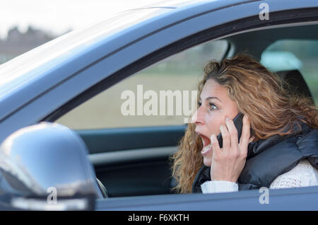 Frau ist ihr Auto mit einem schockiert Ausdruck während eines Anrufs Stockfoto