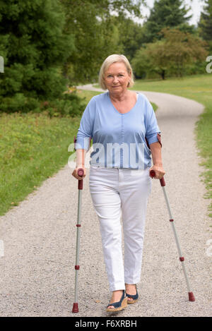 Voller Länge Schuss von einem mittleren Alter Frau zu Fuß auf den Weg mit zwei Stöcken und lächelt in die Kamera. Stockfoto