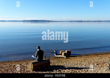 Mann sitzt auf einem Holzscheit mit einem Bier am Rande eines Sees, Feldwieser Bucht, Chiemsee, Oberbayern, Deutschland, Europa. Stockfoto