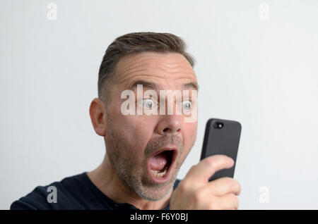 Überrascht, lustigen Mann mittleren Alters mit blauen Augen und einem schockiert Gesichtsausdruck auf den Bildschirm seines Mobiltelefons portra Stockfoto