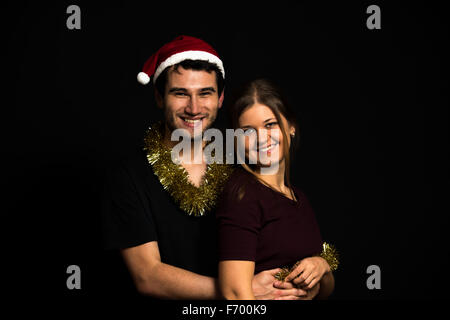 Glücklich lächelnde Paar, tragen Weihnachtsdekoration, umarmen, das Tragen der roten Hütte, auf schwarzem Hintergrund, low-key Stockfoto