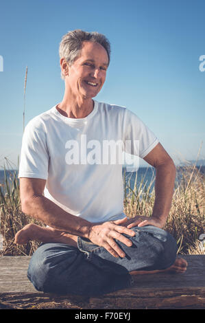 Reifer alter Mann in den sechziger Jahren, der Achtsamkeit, Meditation, Yoga im Freien in der Sonne am Wasser übt. Stockfoto