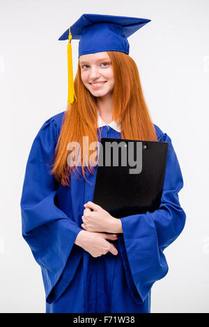 Porträt der hübsche Rothaarige fröhlich studierte positive Mädchen im blauen Kleid und Graduierung Kappe hält Zwischenablage Stockfoto
