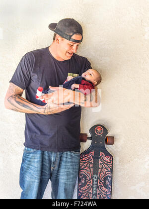Tausendjährige Vater Skateboarding mit Tattoos halten sein kleiner Sohn 2 1/2 Monat und steht neben der Papa Skateboard. Stockfoto