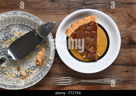 Vogelperspektive Blick auf ein Stück Pecannusskuchen auf einem Teller neben den leeren Kreis Zinn mit Server. Auf einem Holztisch mit Gabel. Stockfoto