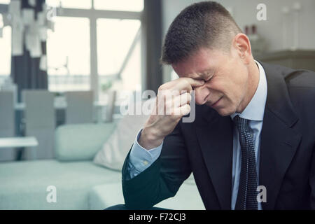 Mann im Anzug sitzt auf der Couch im Wohnzimmer mit Kopfschmerzen Stockfoto