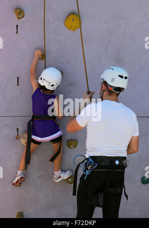 Kinder klettern an einer Kletterwand unter die Überwachung und die Hilfe ihres Vaters während einer lokalen Jugend Sportmesse lernt. Stockfoto