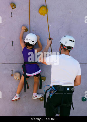 Kinder klettern an einer Kletterwand unter die Überwachung und die Hilfe ihres Vaters während einer lokalen Jugend Sportmesse lernt. Stockfoto