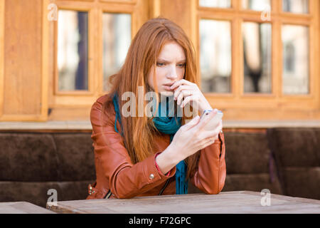 Peinlich, traurig Einschlagloch junge rothaarige Dame lesen Nachricht im Handy im Open-Air-Café sitzen Stockfoto