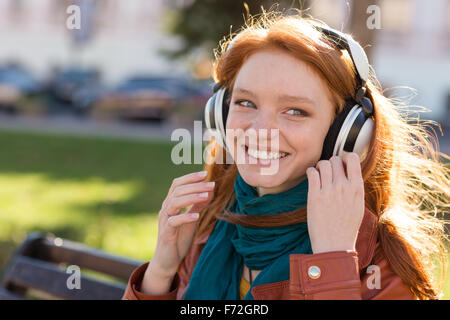 Porträt von glücklich lächelnde rothaarige junge Dame in hellen Schal Musikgenuss auf der Bank im park Stockfoto