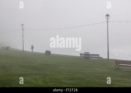 Eine Person, die zwischen Laternenpfosten an einer Strandpromenade im Nebel, Largs, North Ayrshire, Schottland, Großbritannien, läuft Stockfoto