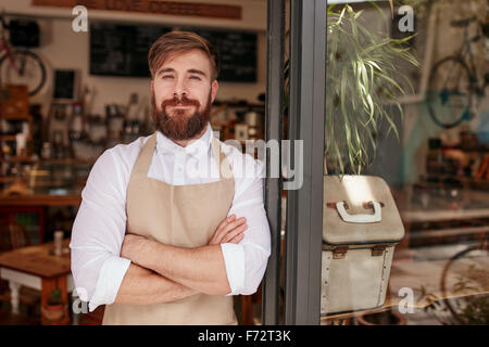 Portrait über eine schöne und selbstbewusste Café-Besitzer stehen vor der Tür. Junger Mann stehend mit seine Arme verschränkt, Blick in die Kamera Stockfoto