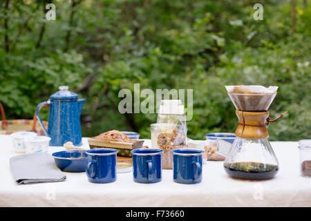 Tabelle in einem Garten, mit einer Kaffeemaschine, Tassen und Schalen, Kuchen und Müsli. Stockfoto