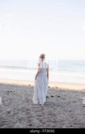 Blonde Frau trägt ein langes Kleid steht an einem Sandstrand. Stockfoto