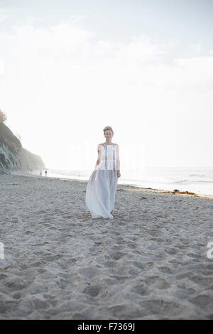 Blonde Frau trägt ein langes Kleid steht an einem Sandstrand. Stockfoto