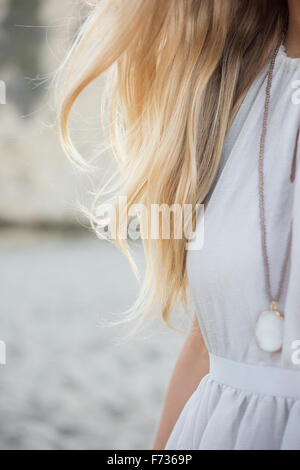 Blonde Frau trägt ein Kleid und Halskette an einem Sandstrand. Stockfoto