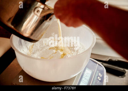 Ein Bäcker Flüssigkeit aus einem kupfernen Krug in einer Schüssel mit Mehl und Zutaten für den Teig gießen. Stockfoto