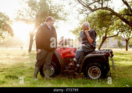 Zwei Männer, ein Bauer und ein Mann mit einem Klemmbrett, durch ein Quadbike in einem Obstgarten. Stockfoto