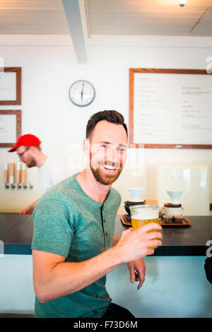 VANCOUVER, BRITISH COLUMBIA, KANADA. Ein junger Mann lächelt mit einem Pint Bier in der Hand, sitzt an der Bar von einem hell erleuchteten Pub. Stockfoto