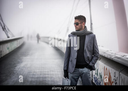 Hübscher trendige junger Mann zu Fuß auf einer Brücke im Winter tragen einen metallischen Aktenkoffer tragen Sonnenbrillen und Schal in einem nebligen Tag Stockfoto