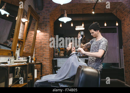 Junge attraktive Friseur mit Tatoo in schwarzen Hut trocknen Mannes Haare mit greift im Friseursalon Stockfoto