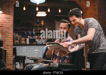 Junge glücklich Barber und gut aussehend zufrieden Kunde sucht durch Magazin sitzen im barbershop Stockfoto