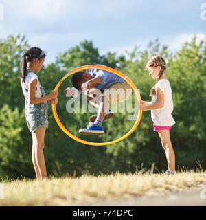 Junge springt durch Hula Hoop Reifen im Park, während seine Freunde es halten Stockfoto