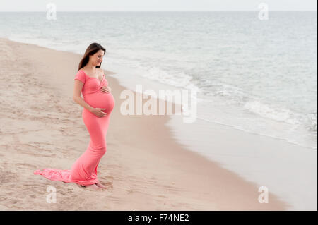 Schöne schwangere Frau eine Koralle gefärbt, volle Länge, Mutterschaft Kleid tragen. Sie lächelt und blickte auf ihren Bauch. Sho Stockfoto