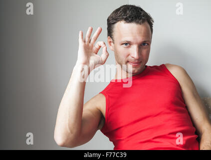 Positiven jungen lächelnder Mann im roten Hemd zeigt Ordnung Geste Stockfoto