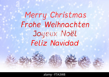"Frohe Weihnachten" geschrieben Englisch, Spanisch, Deutsch und Französisch auf blauem Hintergrund mit Schneeflocken und Zapfen Stockfoto