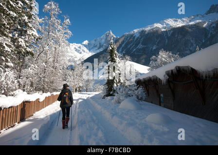 Frau zu Fuß mit Skistöcke schneebedeckten Weg mit Eiszapfen hängen von Dach, bewaldete Hänge und Berggipfel vor uns Stockfoto
