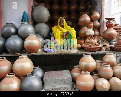 Eine ältere indische Frau in einem hellen gelben und grünen Sari sitzt Schneidersitz unter eine Anzeige von grau - Keramik Terrakotta-Töpfe. Stockfoto