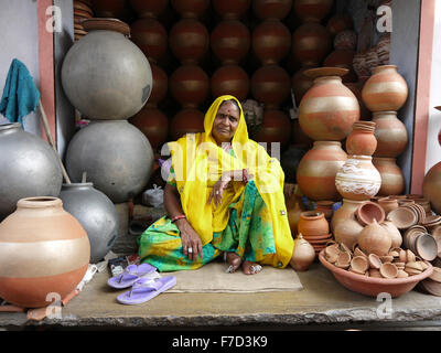 Eine ältere indische Frau in einem hellen gelben und grünen Sari sitzt Schneidersitz unter eine Anzeige von grau - Keramik Terrakotta-Töpfe. Stockfoto