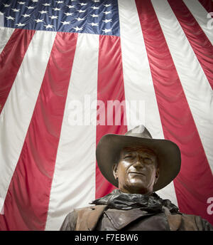 Santa Ana, Kalifornien, USA. 27. November 2015. Eine überlebensgroße Bronzestatue von John Wayne ist auf dem Display an John Wayne International Airport. Wayne, ein Hollywood-Schauspieler berühmt für seine Rollen als harte Cowboys, lebte in Newport Strand. Die Bronzestatue von Wayne wird umrahmt durch eine amerikanische Flagge im Hintergrund hinter zur Veranschaulichung eines Bildes des Patriotismus für eine Generation, die auf seine Filme repräsentieren den heftigen und ikonischen Individualismus viel gehegt und gepflegt von damals in der amerikanischen Geschichte und Kultur aufgewachsen. Wayne ist dargestellt, wie er in den meisten seiner westlichen Filme als robuste Individualist, Hea war, Stockfoto