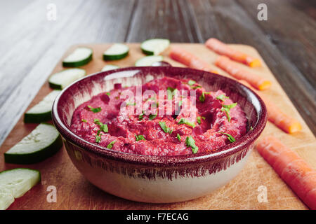 Hummus mit rote Beete in Schüssel auf Schneidebrett, umgeben von Karotten und Gurken auf einem dunklen Holztisch Stockfoto