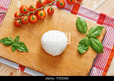 Mozzarella auf Schneidebrett umgeben von Tomaten und Basilikum auf einem Holztisch Stockfoto