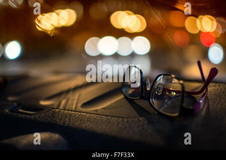 Frauen treibende Brille sitzt auf dem Armaturenbrett eines Autos in der Nacht mit defokussierten Autoscheinwerfer im Hintergrund Stockfoto
