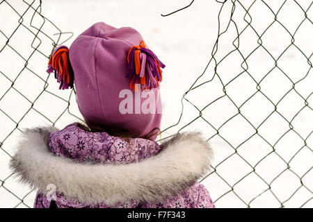 Junges Mädchen in warme Winterkleidung schaut durch einen defekten Maschendrahtzaun Stockfoto