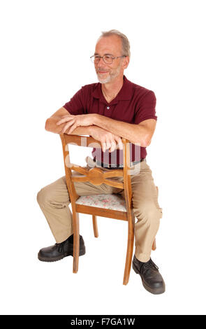 Ein ernster älteren Mann in Beige Hosen und Burgund T-shirt rückwärts auf einem Stuhl sitzend für weißen Hintergrund isoliert. Stockfoto