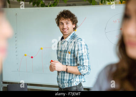 Schöner smart erfolgreich positive lockigen Kerl im karierten Hemd Stand in der Nähe des Whiteboards und lächelnd Stockfoto