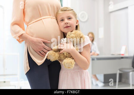 Porträt-Mädchen mit Teddybär umarmt schwangere Mutter in Arztpraxis Stockfoto