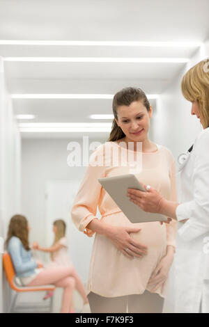 Arzt mit digital-Tablette im Gespräch mit schwangeren Patientin im Klinik-Korridor Stockfoto